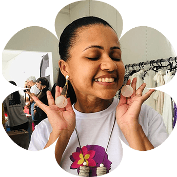 Fabiana Moura, profissional da área da beleza e voluntária do projeto, sorrindo enquanto exibe brincos artesanais, em um ambiente de exposição com roupas ao fundo.